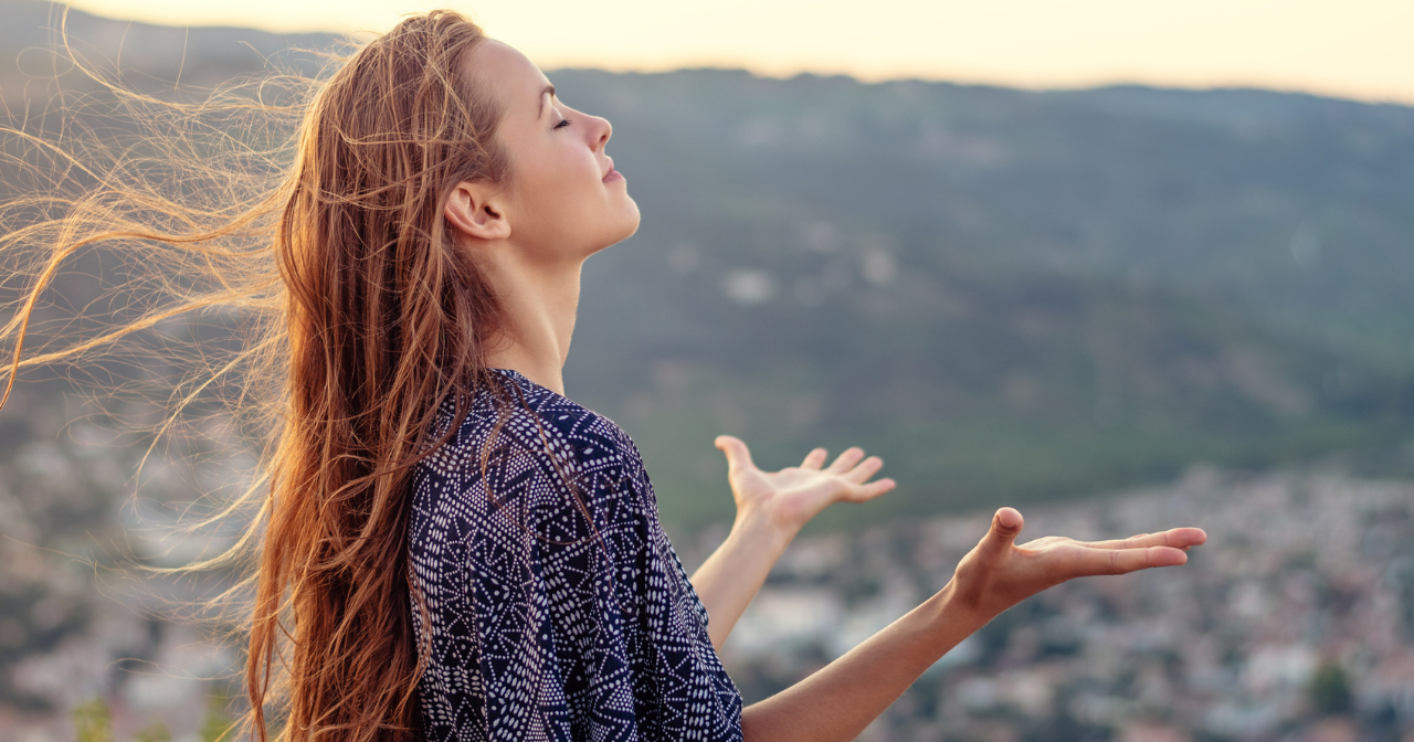 A young woman stands on the top of a hill with her hands outstretched and her face upturned to the sun.