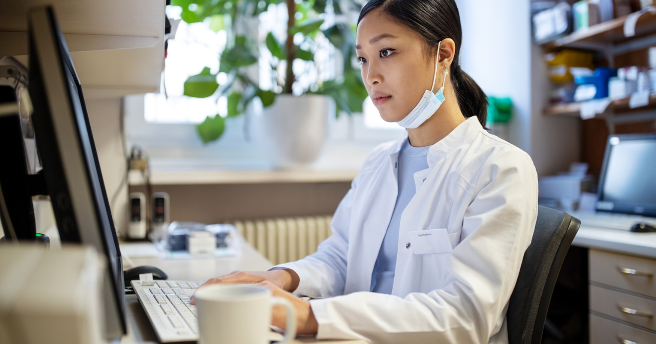 A pharmacist sits alone at a computer with their mask sitting below their face as they work on prescriptions. 