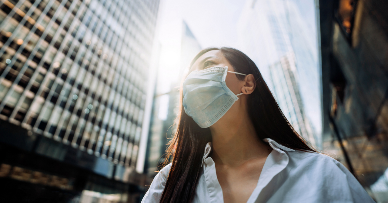 An Asian woman in a mask walks down a city street in business clothes. 