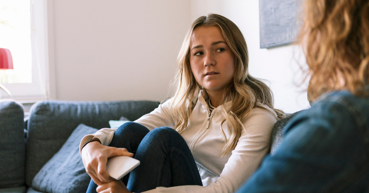 A concerned looking teen hugs her knees to her chest while talking to an adult. 