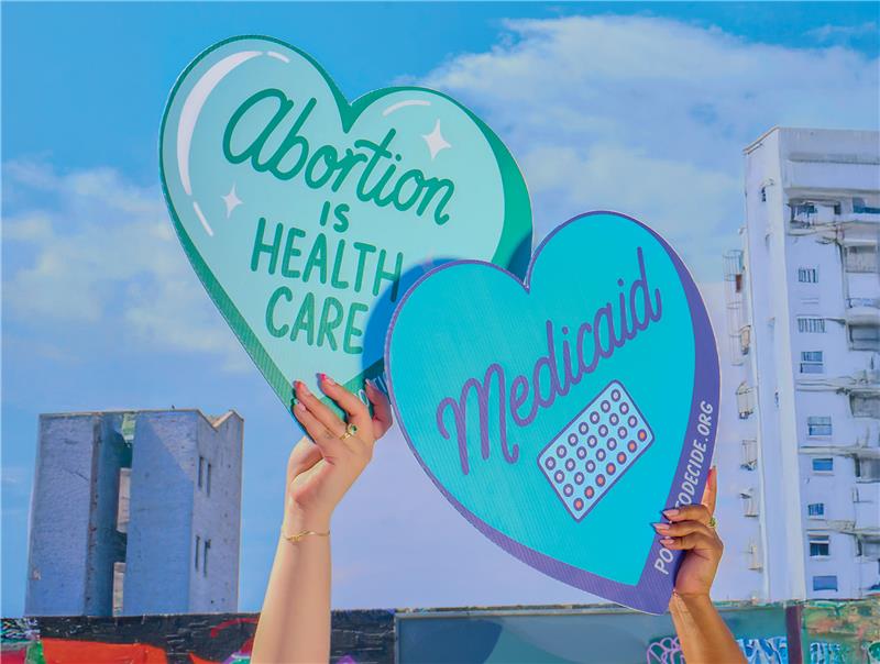 Two hands hold up heart shaped signed. One reads "Abortion is health care" and the other reads "Medicaid."