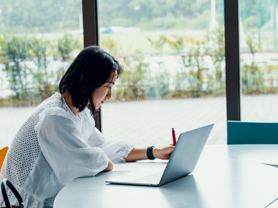 A woman works at her computer while taking notes by hand. 