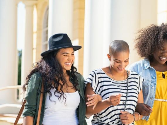 Three young woman link arms and laugh. 