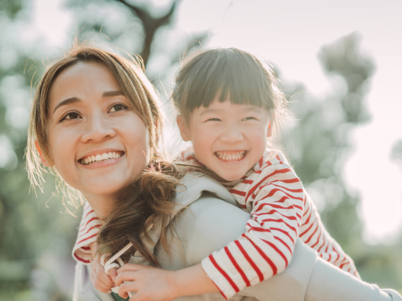 A mother smiles with her young daughter on her back laughing. 