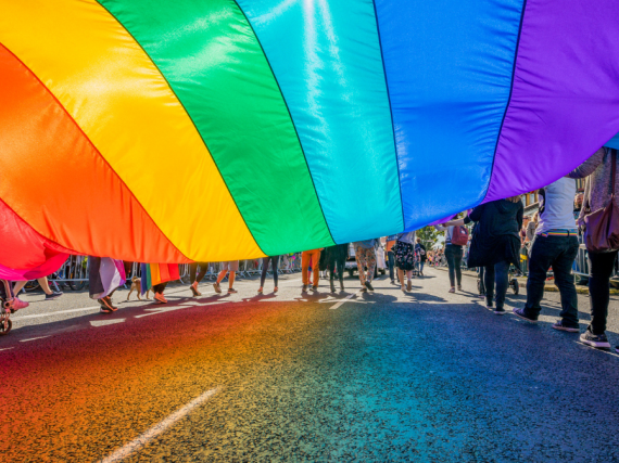 A photo from underneath a massive pride flag being carried by many people during a parade. 