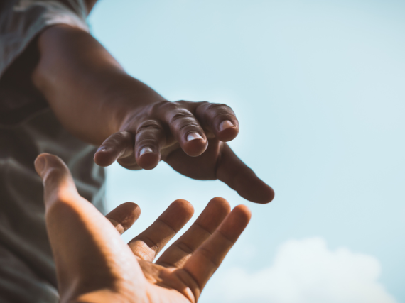 One person's hand reaches down to grasp another person's hands with a blue sky in the background.