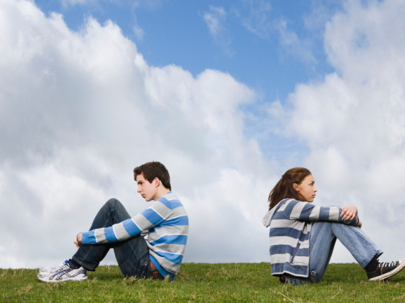 A teen couple sit on the top of a hill hugging their knees with their backs to one another. 