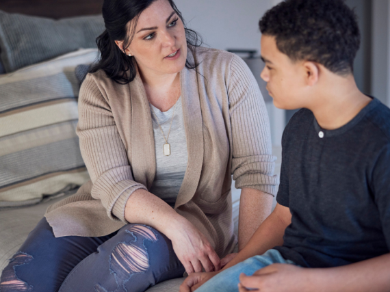 A mom and her son sit on the edge of the bed and talk.