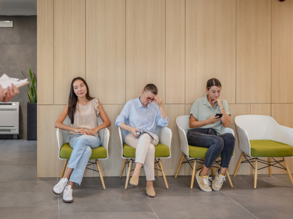 A clinic waiting room with three women waiting in chairs as a health care provider walks past. 